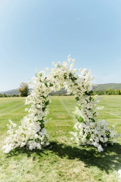 Wedding ceremony arch with white floral arch of roses and hydrangeas, set on a grassy field with distant mountains under blue sky