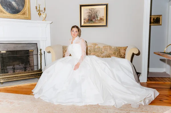 Bridal portrait of a bride in a wedding dress ball gown with lace sleeves, seated on an upholstered sofa before a fireplace and paintings