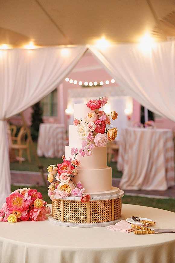 Wedding cake with tiered wedding cake design, cascading sugar and fresh flowers on a rattan stand under draped tent and bistro lights