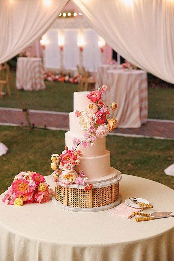 Wedding cake with three tier wedding cake design, smooth white buttercream and cascading pink-coral flowers on a cane stand under string lights