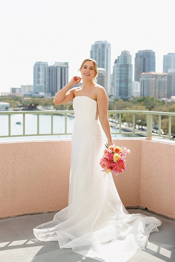 Bridal portrait of a bride in a strapless wedding dress holding a pink-and-orange bouquet on a balcony with river and city skyline behind