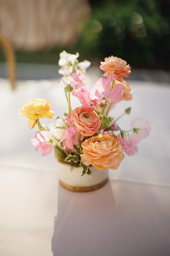 Wedding centerpiece with pastel ranunculus and sweet peas in a white ceramic vase with gold rim on linen, garden chairs softly blurred behind