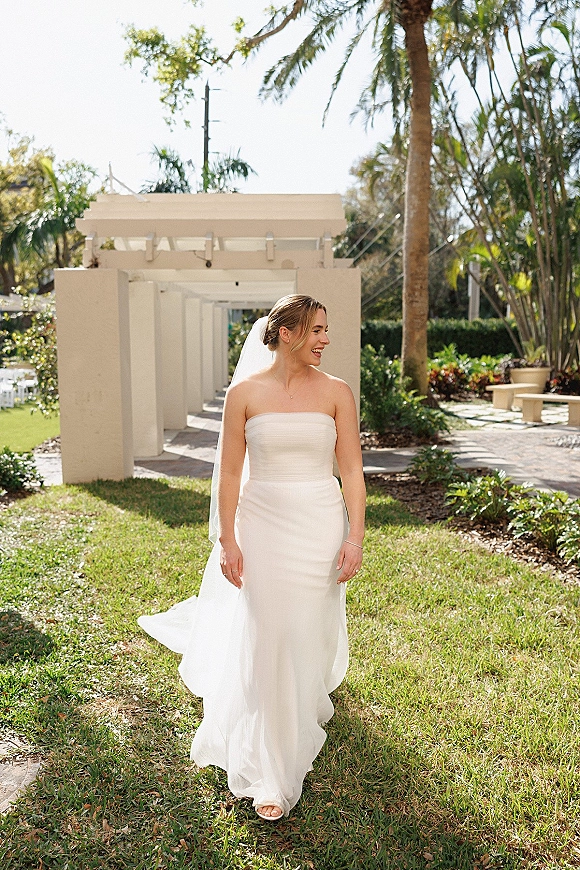 Bridal portrait of a bride in a strapless wedding dress and long veil, showing jewelry and shoes on a garden pergola walkway with palm trees