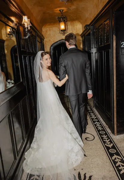 Couple portrait of bride looking back, holding groom’s arm with bouquet and cathedral veil in a moody, dark wood hallway with arched doorway