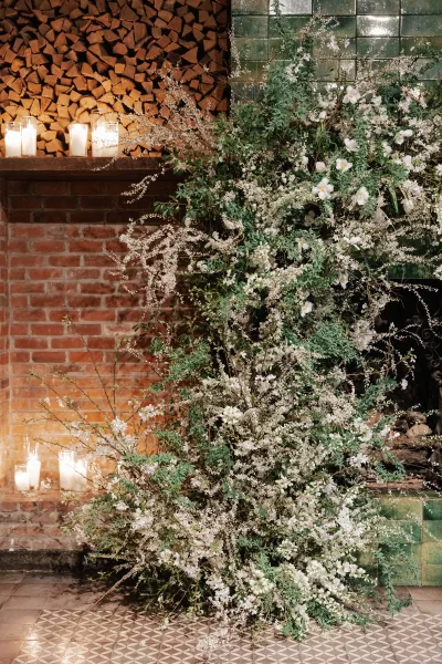 Wedding ceremony decor with a white floral installation and candles on a green tiled fireplace, set against a brick wall and stacked firewood