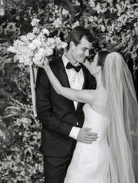 Couple portrait of bride and groom embracing, bride holding a wedding bouquet portrait in front of a lush floral wall backdrop