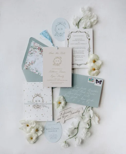 Invitation suite wedding invitation flat lay with dusty blue and sage envelopes, wax seal, tassel, stamps, and white flowers on textured surface
