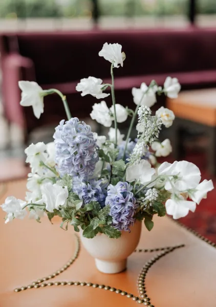 Wedding centerpiece with blue and white flowers and greenery in a ceramic vase, with a beaded garland on a linen table by a window