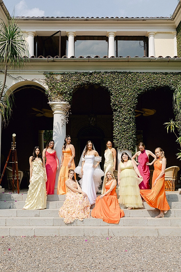 Bridal party portrait of bride with bridesmaids in colorful dresses on stone steps before a columned villa facade under blue sky