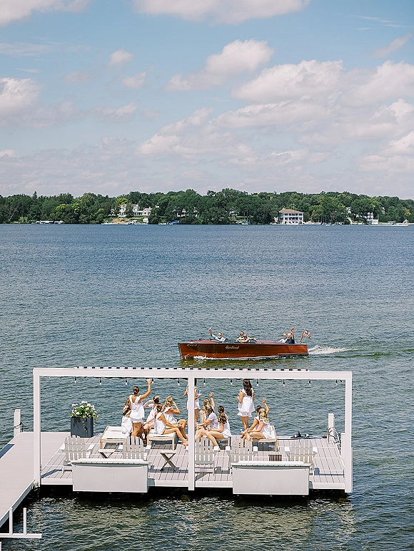 Wedding party boat send off as the bridal party on dock waves beside a classic wooden speedboat, with string lights and lake views under blue sky