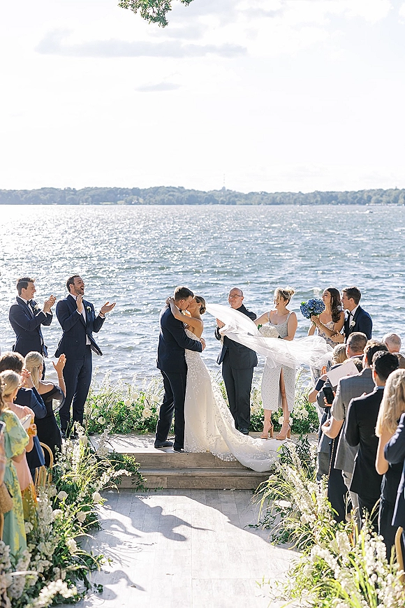 Ceremony kiss at an outdoor wedding ceremony by the lake, bride in veil and gown kissing groom in tux as guests cheer amid aisle florals