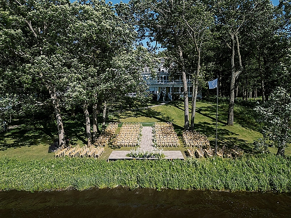 Outdoor ceremony setup with wood chairs flanking an aisle runner, white-green florals, and a platform stage on a riverside lawn under blue sky