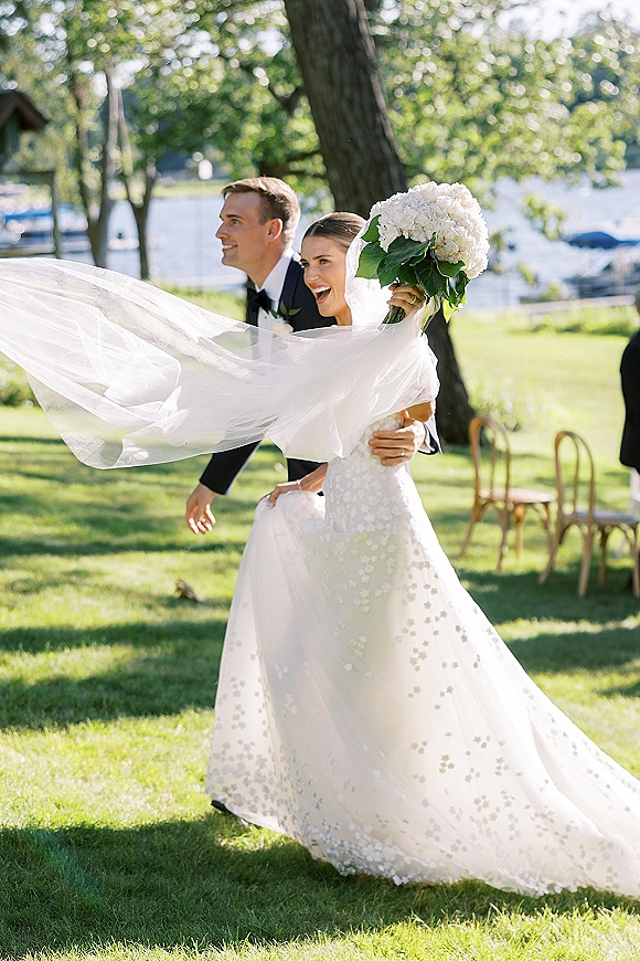 Wedding recessional with bride and groom walking, bride laughing with bouquet and flowing veil beside a lakeside lawn with trees