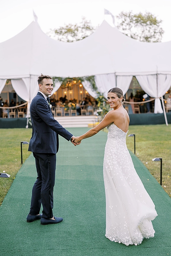 Couple portrait of bride and groom holding hands, looking back under a draped white tent with string lights and greenery garland