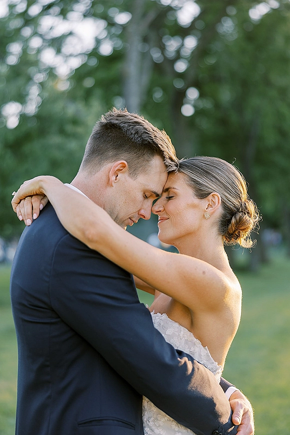 Couple portrait of bride and groom embrace with foreheads touching, eyes closed, in sunlit garden trees, bride in lace dress