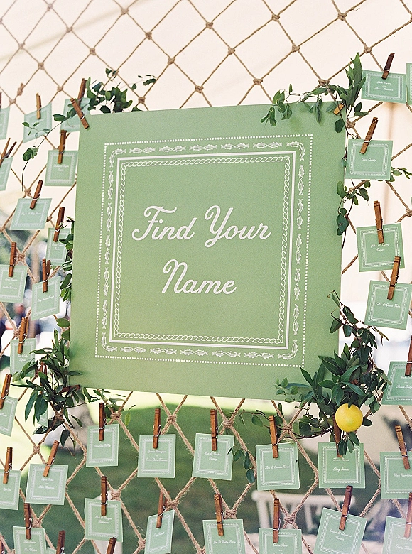 Wedding seating chart with escort card display clipped to a rope net, accented by greenery garland and lemons on an outdoor lawn