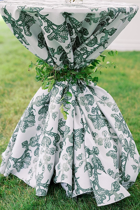 Cocktail table decor on a high top table with patterned botanical linen and a greenery garland, styled for an outdoor lawn cocktail hour
