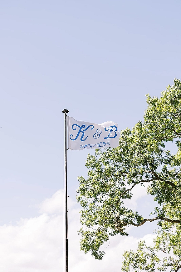 Wedding monogram flag, a custom wedding flag with script initials on a flagpole, fluttering against blue sky with clouds and tree branches