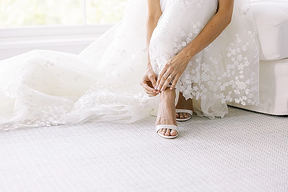 Bride getting ready as she fastens bridal shoes, showing floral lace wedding dress hem, wedding ring, and soft window light by a sofa