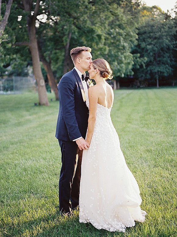 Couple portrait of bride and groom holding hands as he kisses her forehead, her strapless lace gown glowing in golden-hour park light