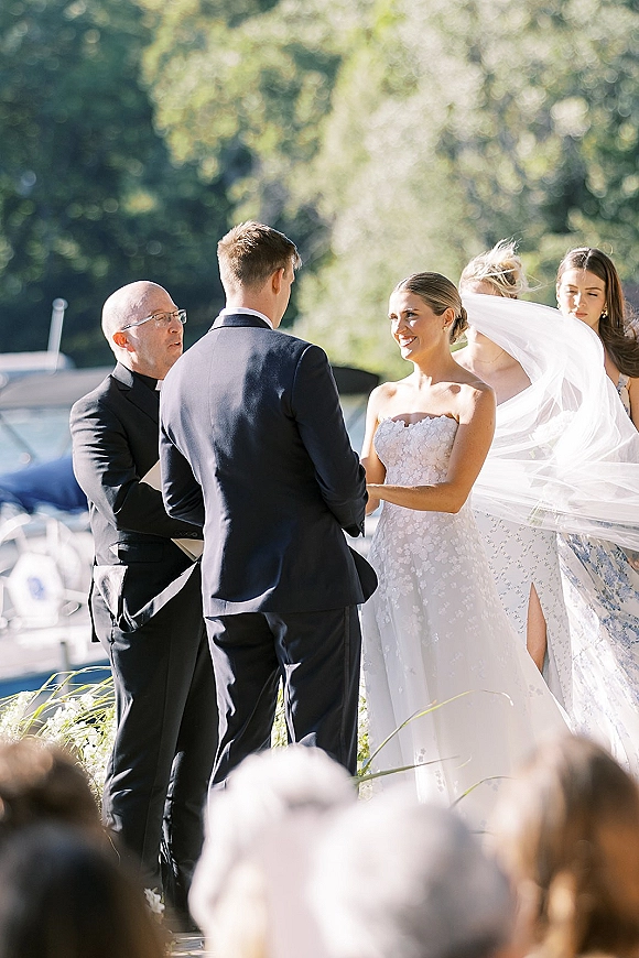 Wedding vows during an outdoor wedding ceremony as bride and groom hold hands by the waterfront, veil blowing, officiant and guests behind