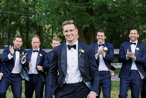 Groom portrait in a navy tuxedo with bow tie and white rose boutonniere, standing on a green lawn with trees and a wooden fence behind