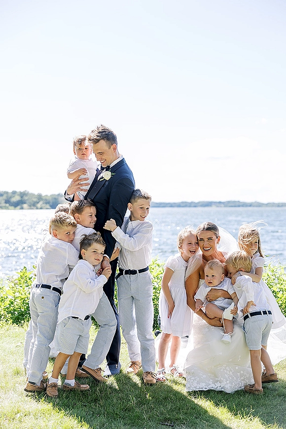 Wedding family portrait of bride with children and groom holding toddler in tux and veil, posed on a lakeside lawn under blue sky