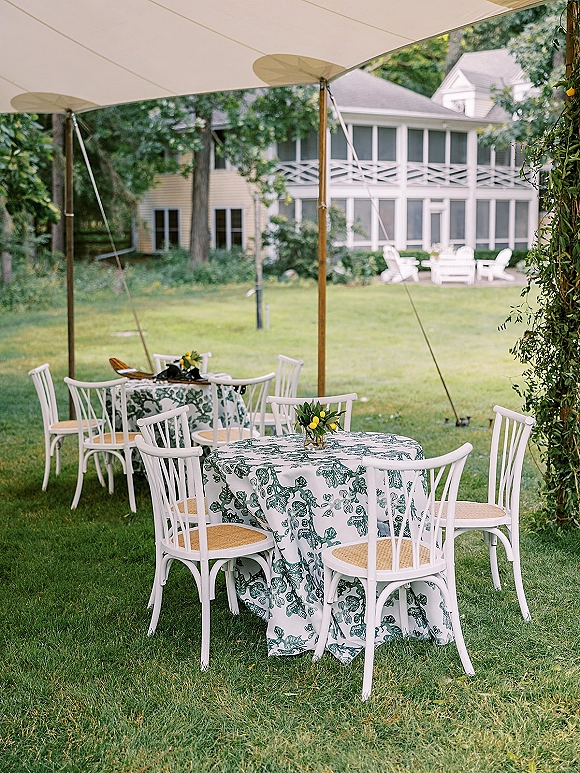 Outdoor reception tables under a sailcloth tent with toile tablecloths, white bistro chairs, lemon florals, and greenery on a lawn