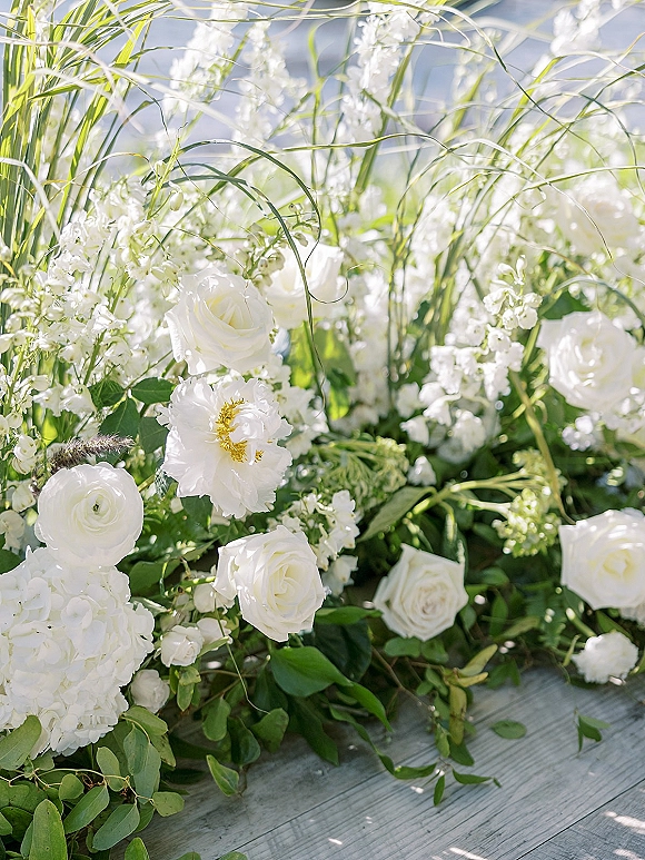 Wedding flowers in a white rose arrangement with ranunculus, stock, eucalyptus, and ornamental grass on a sunlit wood deck floor