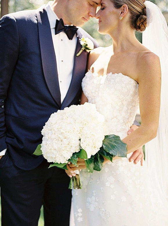 Couple portrait of bride and groom close up, touching foreheads as she holds a white hydrangea bouquet in sunlit greenery bokeh