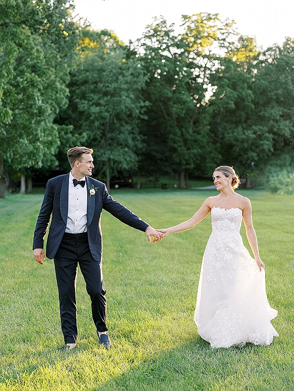 Couple portrait of bride and groom holding hands, walking on a sunlit grassy lawn with trees, her strapless lace wedding dress flowing