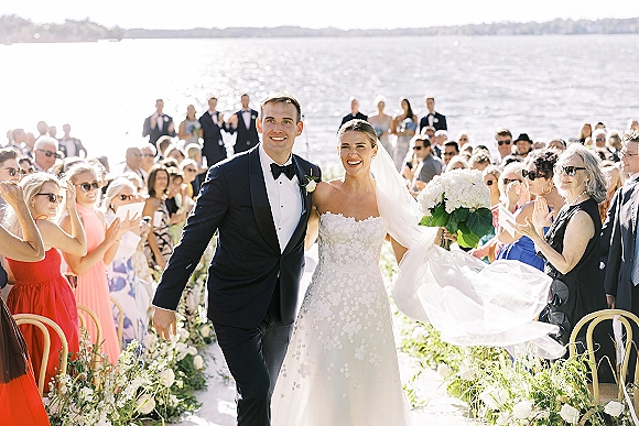 Wedding recessional as bride and groom walk the aisle, her long veil flowing, guests cheering beside waterfront ceremony chairs under blue sky