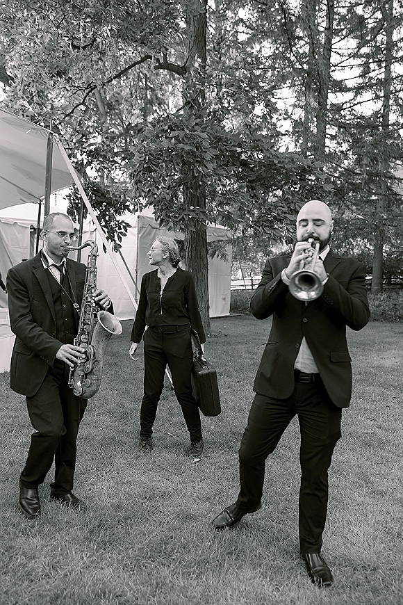 Wedding band in suits with saxophone accent walking across an outdoor lawn near trees and an event tent, playing live music