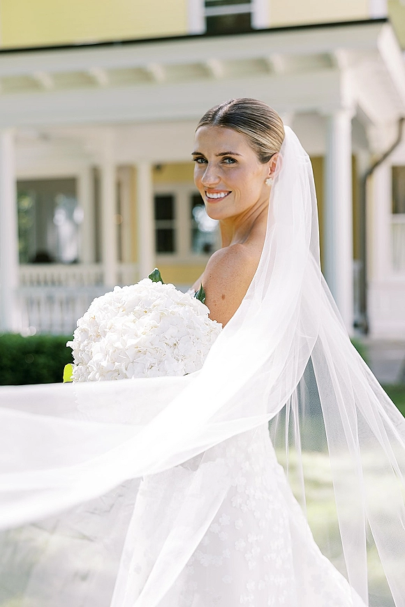 Bridal portrait of a smiling bride with veil blowing, holding a white hydrangea bouquet on a porch with white columns and lawn backdrop