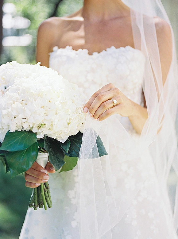 Bridal portrait of a bride holding bouquet of white hydrangeas, veil over a strapless dress with floral appliqués in window light greenery bokeh