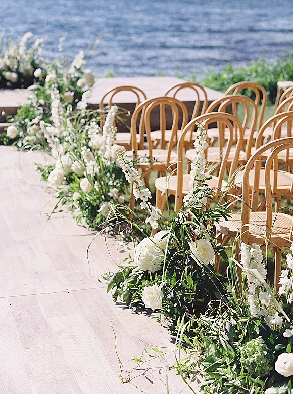 Ceremony aisle decor with wedding aisle flowers, white blooms and greenery garland beside wooden chairs on a wood runner by the waterfront lawn