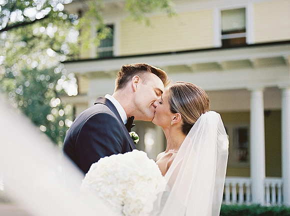 Wedding kiss portrait of bride and groom kissing, bridal veil and white bouquet in foreground, outside a white house porch with columns