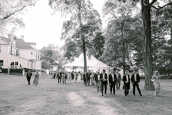 Wedding guests in formal suits and long dresses walk with drink cups toward a white tent reception on a sunny lawn with trees