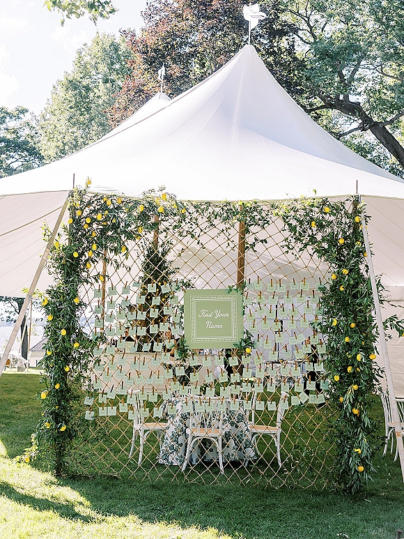 Seating chart display with wedding escort card display on a lattice wall, greenery garland and lemon accents under a daylight tent canopy
