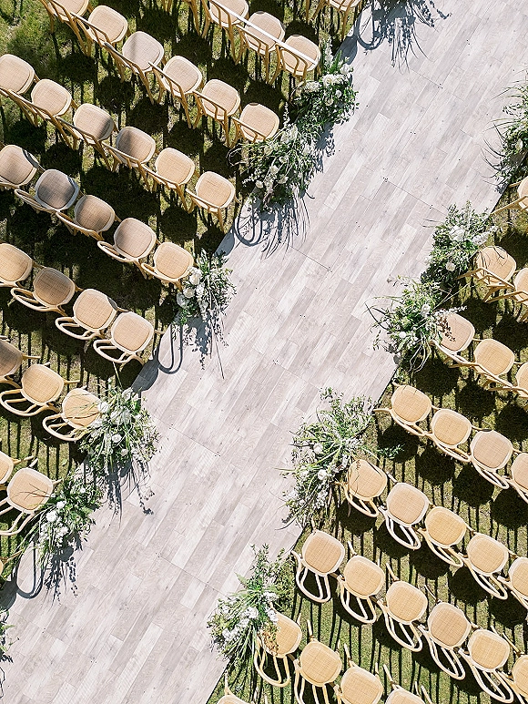 Ceremony aisle design with outdoor ceremony aisle runner, light wood chairs and white florals with greenery on a sunlit lawn
