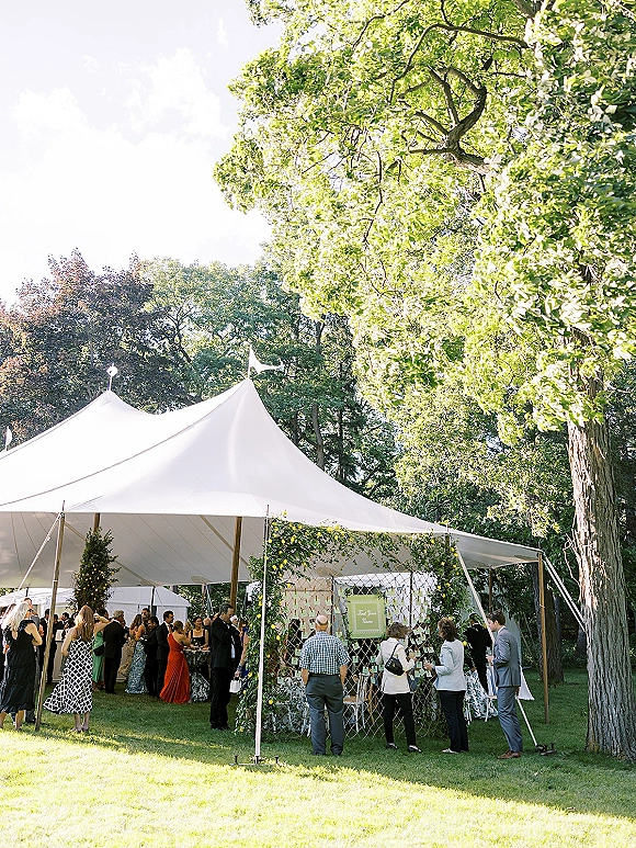 Wedding reception tent with sailcloth canopy and string lights, escort card display and greenery garland on a lawn beneath tall trees
