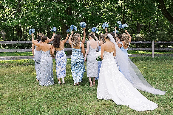 Bridesmaid group photo of bride with bridesmaids from behind, holding blue hydrangea bouquets on a lawn by a wood fence