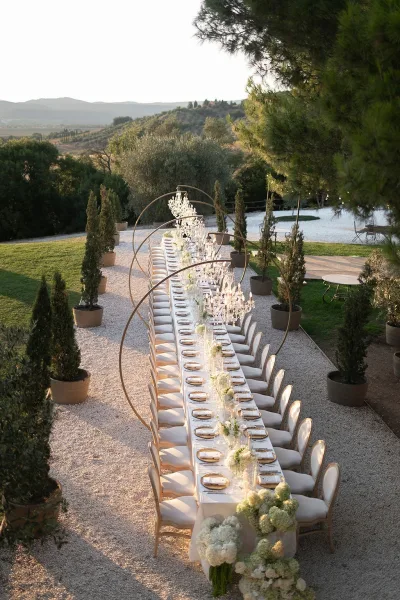 Reception tablescape with a long banquet table setup, gold-rim plates, taper candles, and hydrangeas beneath crystal chandeliers on a gravel patio with mountains beyond