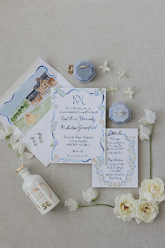 Wedding invitation suite with blue floral wedding invitations, monogram cards, rings in velvet boxes, perfume and white blooms on linen backdrop