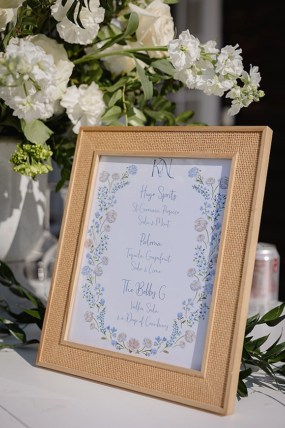 Wedding bar menu framed in burlap with watercolor blue florals and monogram initials on a white tabletop in soft outdoor light