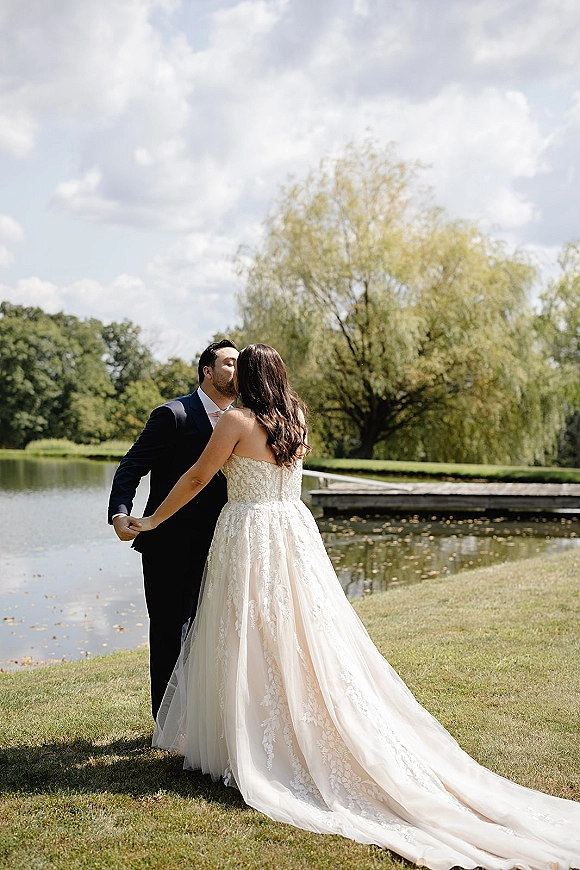 Wedding kiss portrait of bride and groom kissing, holding hands on a wooden dock by a lake, her strapless lace dress train flowing behind