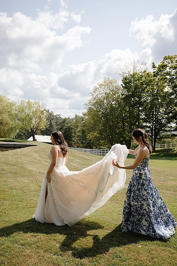 Bride getting ready in a strapless lace wedding dress as a bridesmaid fluffs her long train on a lawn with trees and white fence