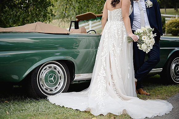 Couple portrait of bride and groom by car, bride in strapless lace dress holding white rose bouquet beside a vintage convertible on a lawn