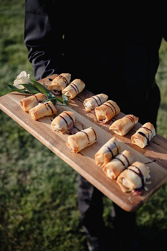 Wedding appetizers on a rustic serving board with bite-size pastries, balsamic drizzle, and greenery garnish held by a server on a lawn