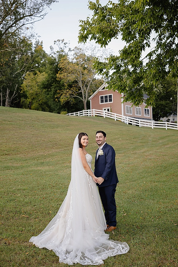 Couple portrait of bride and groom holding hands, her veil and lace train flowing beside his navy suit on a grassy hill by a farmhouse fence